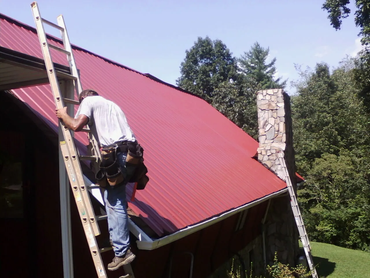 Roofer on a ladder beside a red metal roof and stone chimney — Soffit & Fascia Repair by Roofing Brothers in Rappahannock Academy